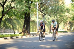 Two men cycle on a quiet road by a park on a summer's day.
