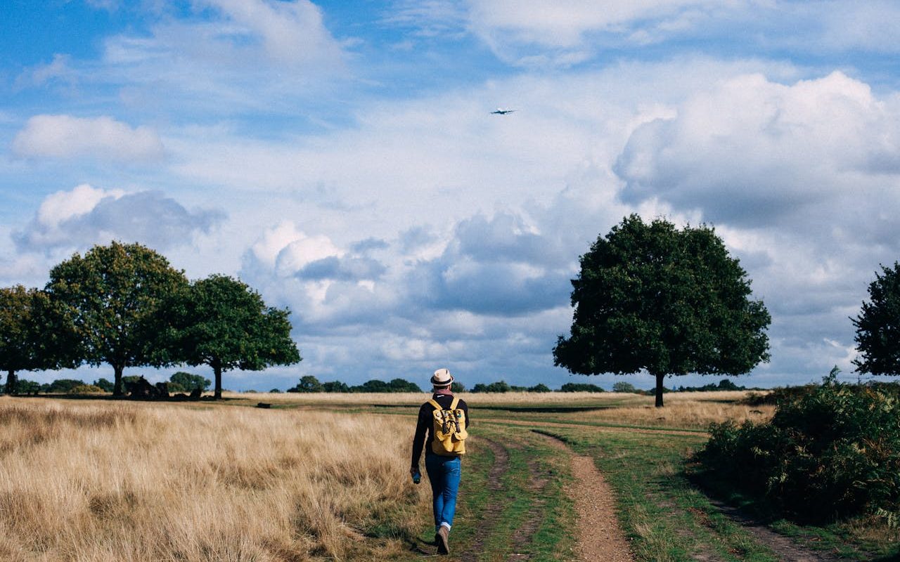 A man walks through the countryside on a sunny day. He wears a hat and carries a rucksack.