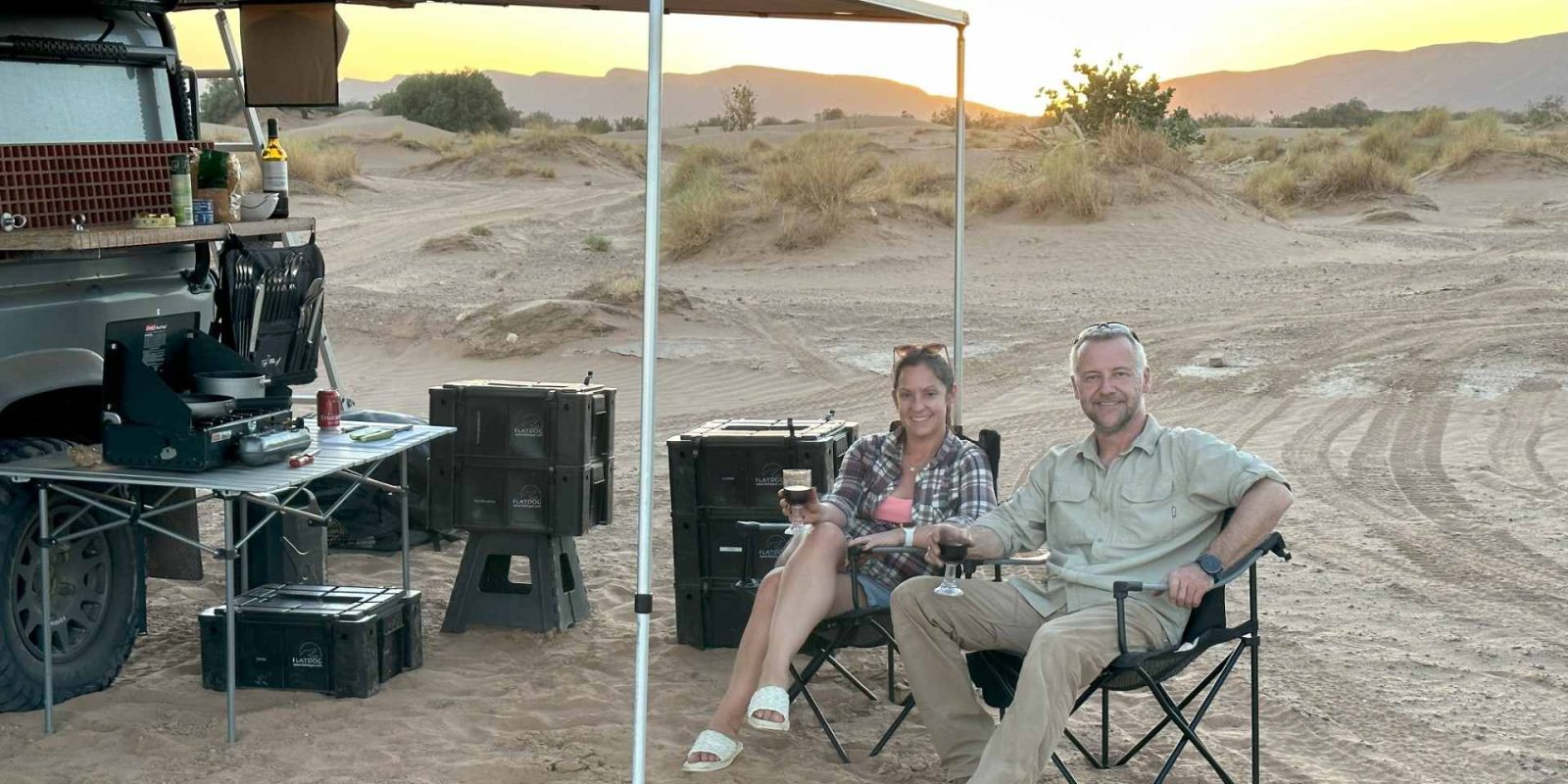 A male and female sit under the awning of their campervan setup in a desert landscape.