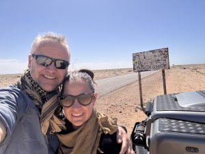 Adventurer James Whitford and partner Emma in sunglasses, hugging. There's a Tropic of Cancer road sign in the background in the Sahara desert.