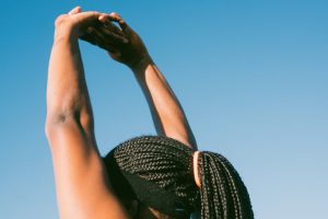 A Black woman stretches her arms to the blue sky. She is wearing a black headband over her braids. Her face is hidden.