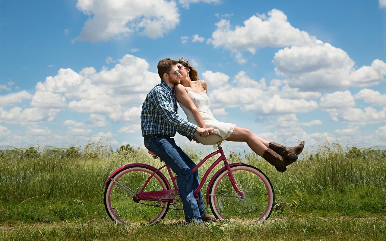 Couple riding he same bicycle across a field - man astride and woman on the handlebars.