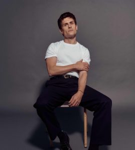 Singer-songwriter Tom Speight sits on a chair in white T shirt and black trousers, against a grey background.