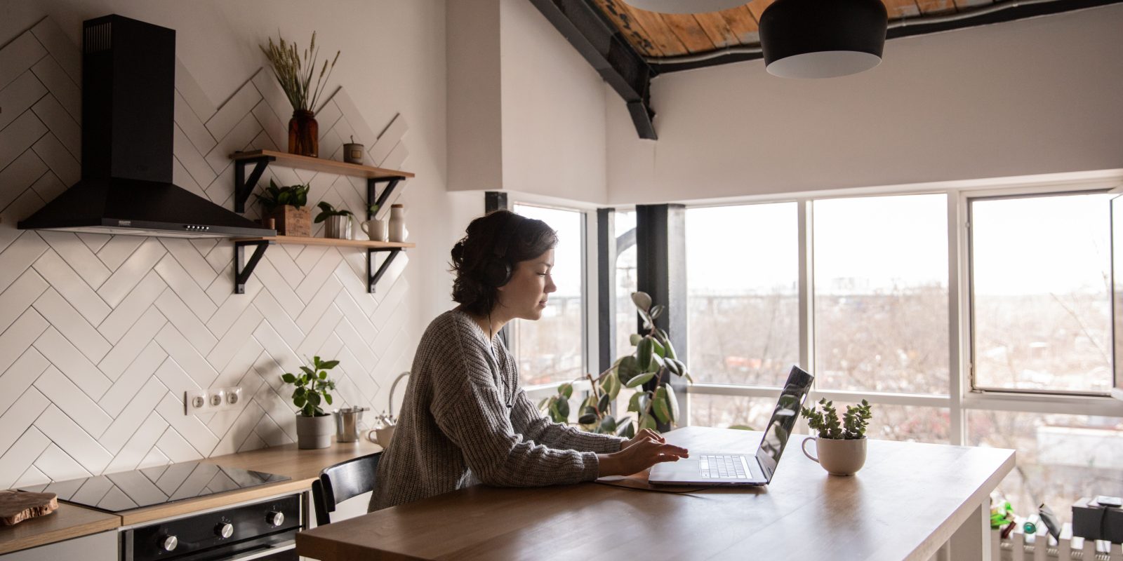 A young woman works on her laptop in a modern, bright kitchen setting with natural decor.