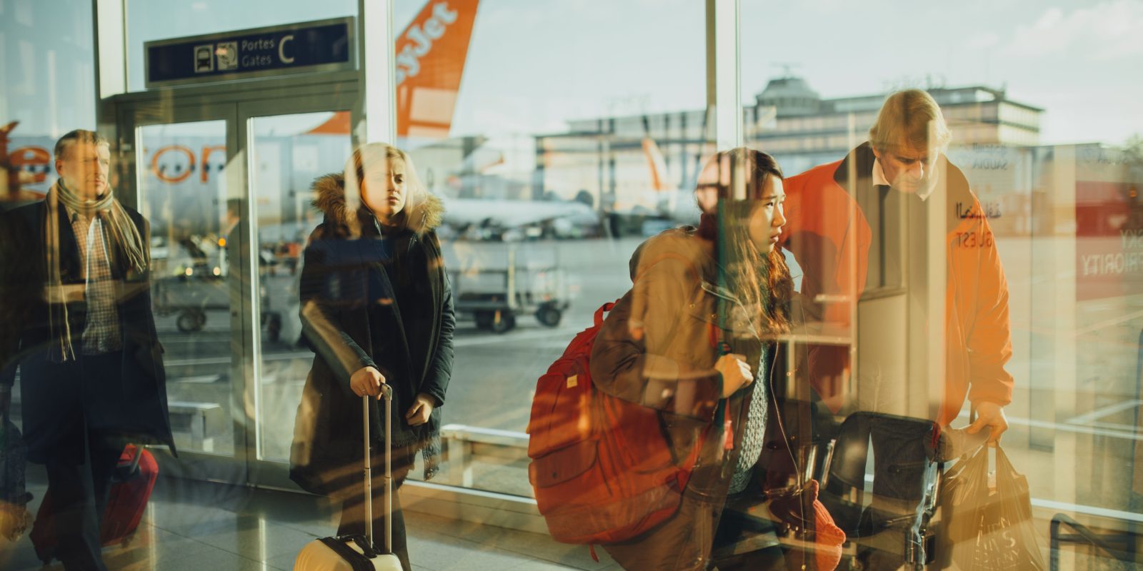 Passengers walking through an airport terminal corridor with large windows overlooking aircraft on the runway.